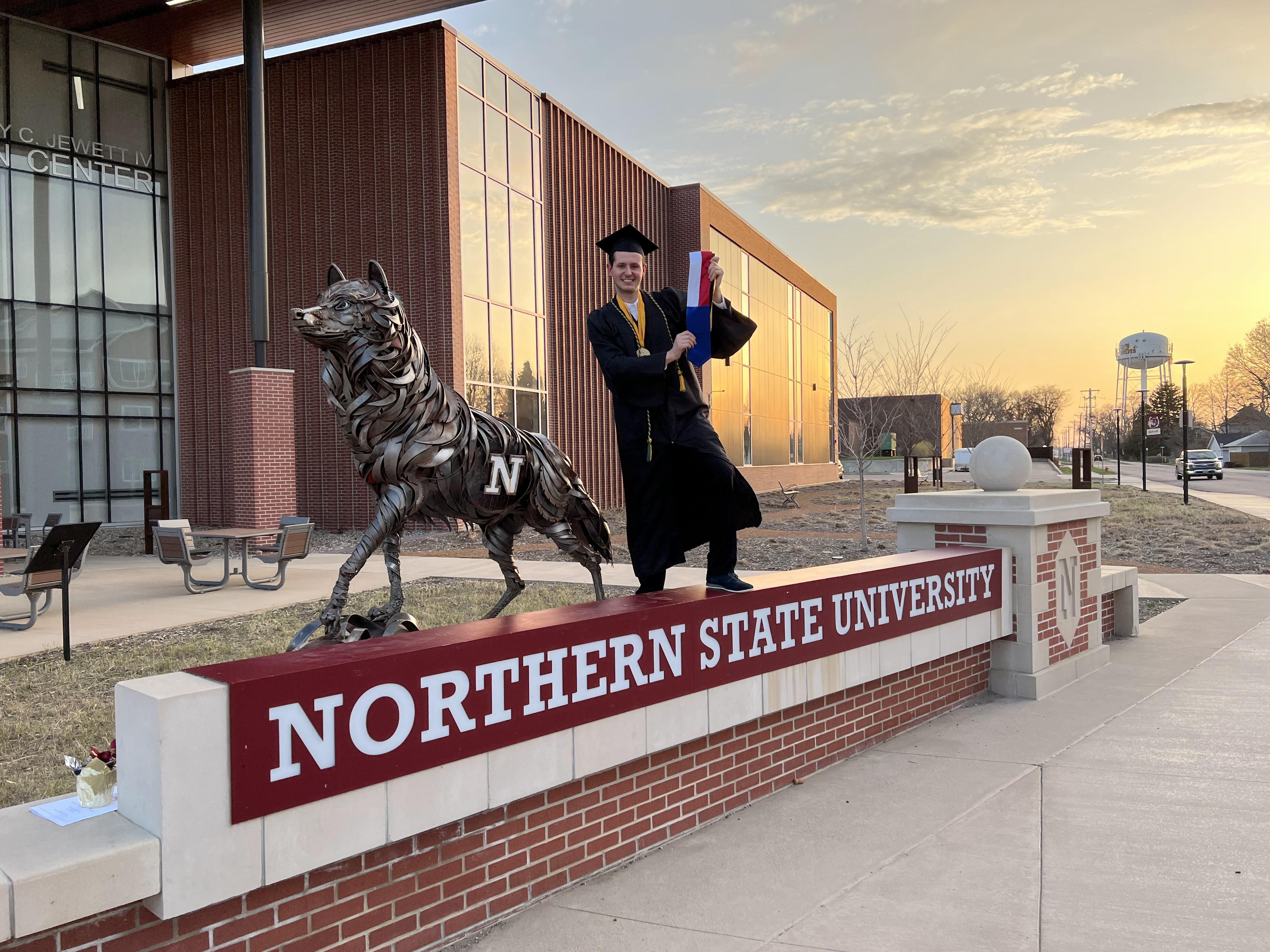 Alum Matj ?p in cap and gown standing by the NSU Wolf statue